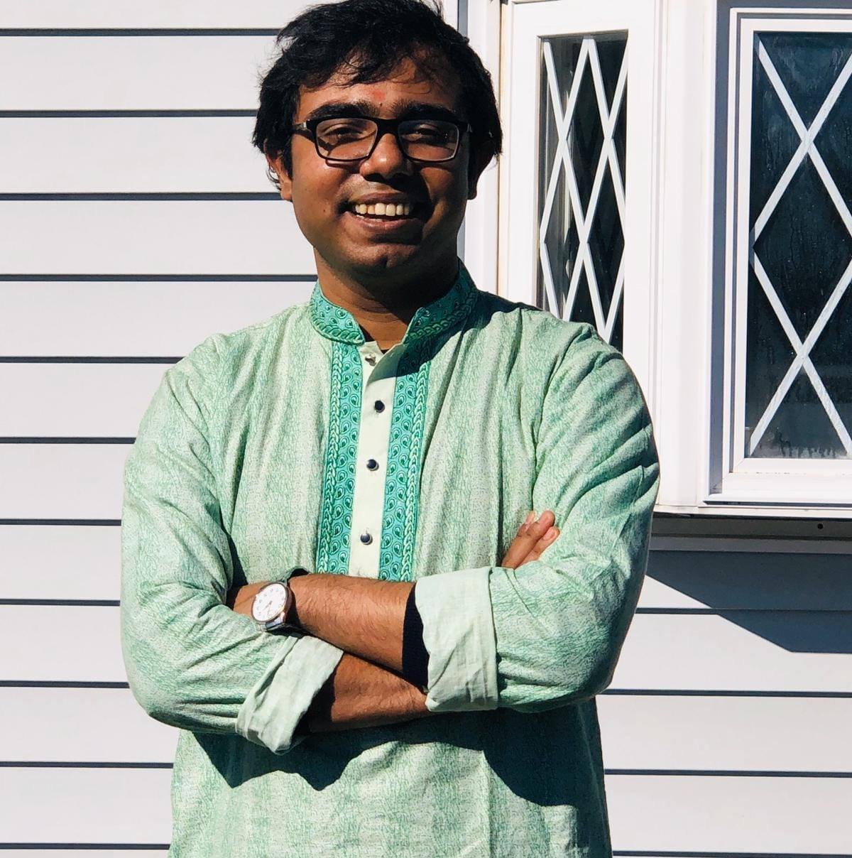 Dipto, a Brown man with black straight hair, is smiling at the camera. She is wearing square glasses and a light green long vest and standing in front of a white wooden wall.